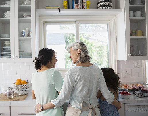 Grandmother, daughter, and granddaughter standing in a kitchen, talking and laughing at the sink